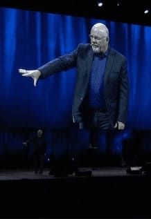 A man is speaking from a stage and is projected onto a big screen.